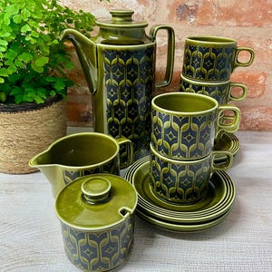 May include: A ceramic tea set in olive green featuring a dark geometric pattern. The set includes a tall teapot with a lid, a creamer, a lidded sugar bowl, and four stacked cups with matching saucers, all decorated with a repeating stylised motif.