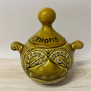 May include: A glazed olive green ceramic onion jar with a lid featuring a spherical knob. The jar has two side handles and a moulded face design with prominent eyes and a nose. The word Onions is written in black script on the lid surface.