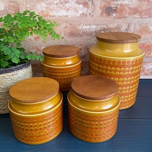 May include: Set of four vintage ceramic canisters with wooden lids. The canisters are brown and orange with a repeating pattern of circles. The canisters are labelled "Coffee", "Sugar", "Tea", and "Biscuits".
