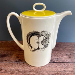 May include: A white ceramic coffee pot with a yellow lid and a black and white illustration of a strawberry on the side. The pot has a handle and a spout.