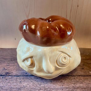 May include: A ceramic cookie jar with a whimsical design. The jar features a light beige face with carved eyes, a nose, and a smiling mouth. The lid is a glossy brown, donut-shaped piece. The jar is set against a wooden background.
