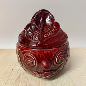 May include: A dark red glazed ceramic jar with a lid shaped like a cabbage leaf. The lid features the embossed text "PICKLED CABBAGE". The base of the jar is moulded with a stylised face featuring circular eyes, a round nose, and a curved mouth.
