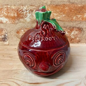 May include: A ceramic beetroot-shaped jar with a lid. The jar is a deep red colour with the word "BEETROOT" embossed on the front. The lid has green leaves and a stem. The jar has a smiling face design.