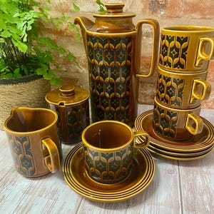 May include: A brown ceramic coffee set with a black and gold geometric design. The set includes a coffee pot, creamer, sugar bowl, cups, and saucers. The cups are stacked, and the set is arranged on a light wood surface.