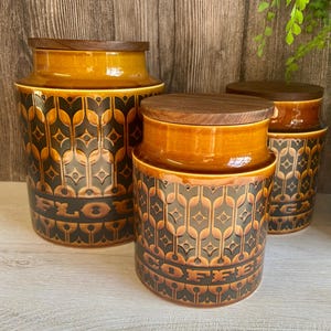 May include: Three brown ceramic canisters with wooden lids. The canisters have a geometric pattern and the words "Flour", "Coffee", and "Sugar" are printed on the front.