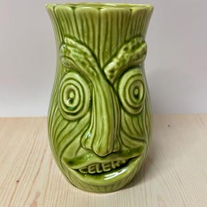 May include: A light green ceramic tiki mug shaped like a stalk of celery with a textured surface, a prominent nose, spiral eyes, and a wide mouth. The word CELERY is embossed in capital letters on the lower lip of the mug, which has a glossy finish.