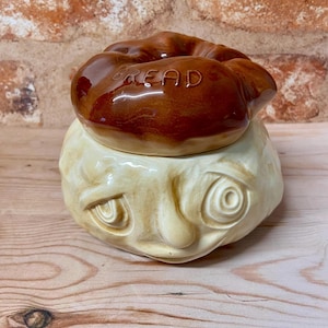May include: A ceramic bread jar with a whimsical face design. The jar is cream-coloured with a brown lid. The lid is shaped like a loaf of bread and has the word "BREAD" embossed on top. The jar has a unique, artistic appearance.