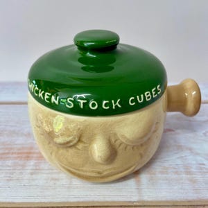 May include: A ceramic storage jar with a beige base shaped like a face with closed eyes and a smile. It features a glossy green lid with the text CHICKEN STOCK CUBES embossed in white lettering and a small handle extending from the side.