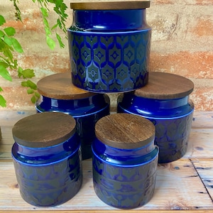 May include: Five blue ceramic storage jars with wooden lids. The jars feature a geometric pattern in a darker blue and are arranged on a wooden surface. The lids are round and made of light brown wood. The jars are suitable for kitchen storage.