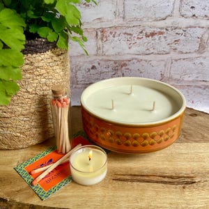 May include: A large, round candle with three wicks in a brown ceramic bowl with a floral design. A small lit candle, matches, and a fern plant are on a wooden surface. The text "BIZARRE" is visible.