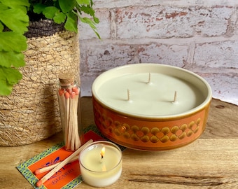 May include: A large, round candle with three wicks sits on a wooden surface. The candle is in a brown ceramic bowl with a floral design. A small lit candle, matches, and a fern plant are also present.