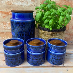 May include: A set of blue ceramic kitchen storage jars with wooden lids. The jars are labelled with the words "Sugar", "Ginger", "Marjoram", and "Allspice". A woven basket with a basil plant is also visible.