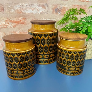 May include: Set of three brown ceramic canisters with wooden lids. The canisters have a black and gold geometric pattern and the words "Sugar", "Flour", and "Tea" are printed on the front.