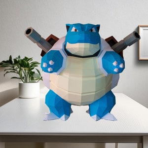 Könnte beinhalten: Ein blaues und weißes Origami-Papiermodell eines schildkrötenartigen Pokemon-Charakters mit Kanonen auf dem Rücken. Das Modell steht auf einem weißen Tisch.