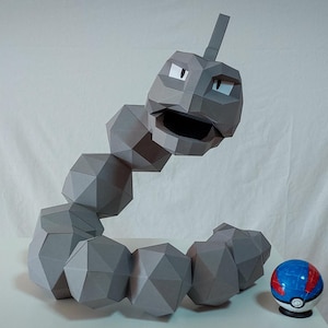 Pode incluir: Uma escultura de papel cinza de Onix, um Pokémon, com um corpo segmentado e uma cabeça grande. A escultura está posicionada ao lado de uma Poké Bola azul e vermelha. O fundo é uma superfície branca lisa.