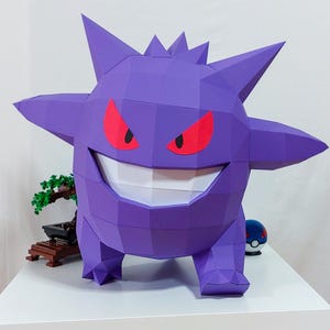 Könnte beinhalten: Eine große, lila Papierfigur von Gengar, einem Pokémon-Charakter, mit roten Augen und einem breiten Grinsen. Die Figur steht auf einer weißen Oberfläche, mit einem Bonsai-Baum und einem Pokéball im Hintergrund.