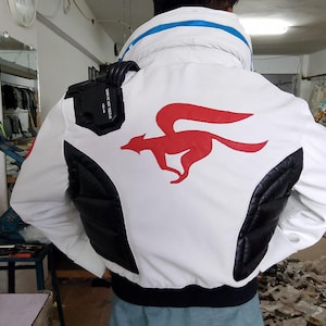 Könnte beinhalten: Weiße Jacke mit roter Fuchsgrafik auf dem Rücken. Die Jacke hat schwarze, gesteppte Einsätze an den Seiten und einen blauen Streifen am Kragen. Eine schwarze Tasche ist an der Schulter befestigt.
