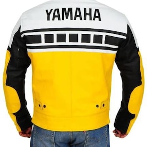 Op de afbeelding: Een gele, zwarte en witte leren jas met het woord "YAMAHA" op de rug gedrukt. De jas heeft zwarte mouwen en een zwart-wit ontwerp over de bovenrug. Het onderste deel van de jas is geel.