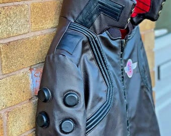 May include: A brown leather jacket with black accents and a red lining. The jacket has a zipper closure and a patch on the left side. The jacket has three black buttons on the left sleeve.