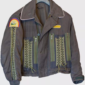 May include: A dark gray jacket with a white collar and green zipper details. The jacket has a rainbow patch on the left sleeve and a yellow patch on the right side. The jacket is decorated with green laces.