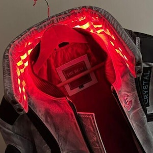 Puede incluir: Una chaqueta gris con forro rojo y un parche negro que dice "CRYSTAL". La chaqueta tiene una cremallera y un cuello. El forro está iluminado con luces rojas.