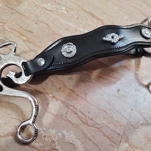 May include: A silver horse bit with a black leather strap and silver accents. The strap has a decorative design with a floral pattern.