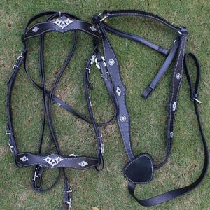 Puede incluir: Bridón de caballo de cuero negro con detalles plateados y costuras. El bridón incluye un frontal, un bozal, riendas y una barboqueja.