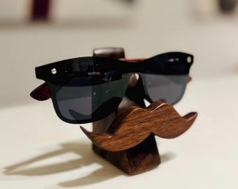 May include: A wooden mustache-shaped stand with a pair of black sunglasses resting on top. The stand is made of dark wood and has a natural finish.