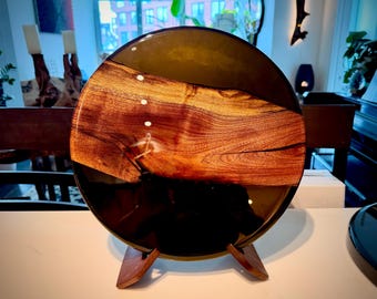 May include: A circular decorative board featuring a central horizontal slice of natural wood with prominent grain patterns, encased in a smooth, dark, translucent resin. The piece is supported by a small, two-piece wooden stand.