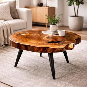 May include: A round coffee table featuring a natural wood slab top with a polished finish, visible wood grain, and a dark central void. The table is supported by three tapered black metal legs and sits on a light-colored textured area rug.