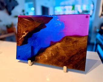 May include: A rectangular serving board made of natural wood with a live edge, featuring a vibrant inlay of translucent purple and deep blue epoxy resin. The resin creates a swirling, river-like effect between the two distinct wooden sections.