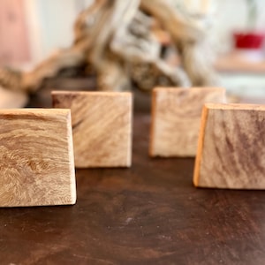 Op de afbeelding: Vier vierkante houten onderzetters met een natuurlijk houtnerfpatroon. De onderzetters zijn lichtbruin met donkerdere bruine strepen en zijn gerangschikt op een donkerbruin oppervlak. De onderzetters zijn waarschijnlijk voor gebruik met dranken.