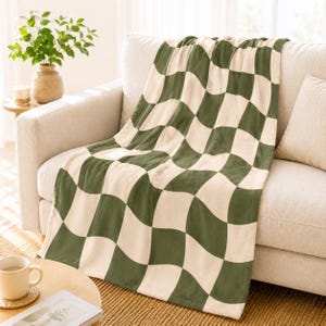 May include: A soft, green and cream checkered blanket draped over a beige sofa. The blanket's pattern features large, wavy squares. The sofa is in a well-lit room, with a small table and a plant in the background. The blanket is a cozy home decor item.