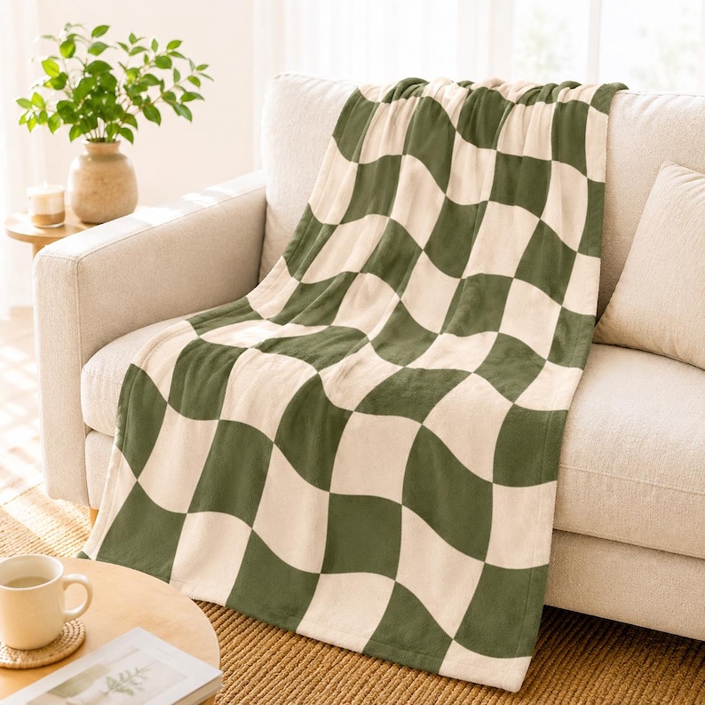 May include: A soft, green and cream checkered blanket draped over a beige sofa. The blanket's pattern features large, wavy squares. The sofa is in a well-lit room, with a small table and a plant in the background. The blanket is a cozy home decor item.