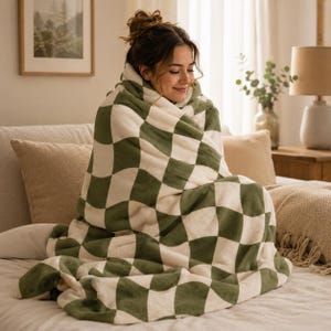 May include: A cozy, oversized blanket with a checkerboard pattern in olive green and cream. The plush blanket is wrapped around a person sitting on a bed, creating a warm and inviting scene. The blanket appears soft and comfortable.