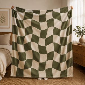 May include: A large, soft blanket with a wavy checkerboard pattern in olive green and cream. The blanket is held up, showcasing its size and design. The cozy throw blanket is perfect for a bed or sofa.