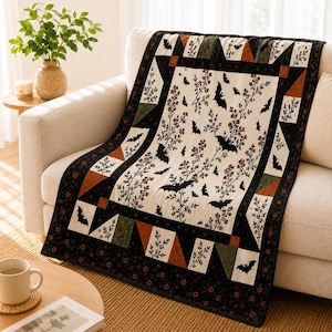 May include: A rectangular quilt featuring a cream-coloured centre panel with a repeating pattern of black bats and brown leafy branches. The border consists of black fabric with small orange flowers, accented by triangular patches in orange and olive green.