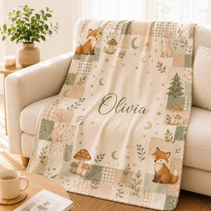 Peut inclure: Un plaid de couleur crème avec une bordure style patchwork ornée d'illustrations de renards, de lapins, de champignons, de sapins et de croissants de lune. Le centre du plaid affiche le prénom Olivia en écriture cursive foncée.