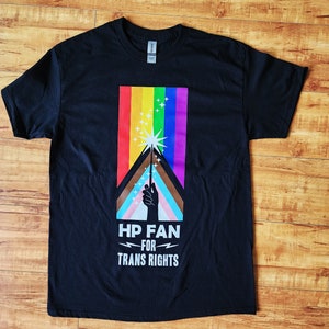 May include: Black t-shirt featuring a rainbow and a hand holding a wand. The text on the shirt reads "HP Fan for Trans Rights".