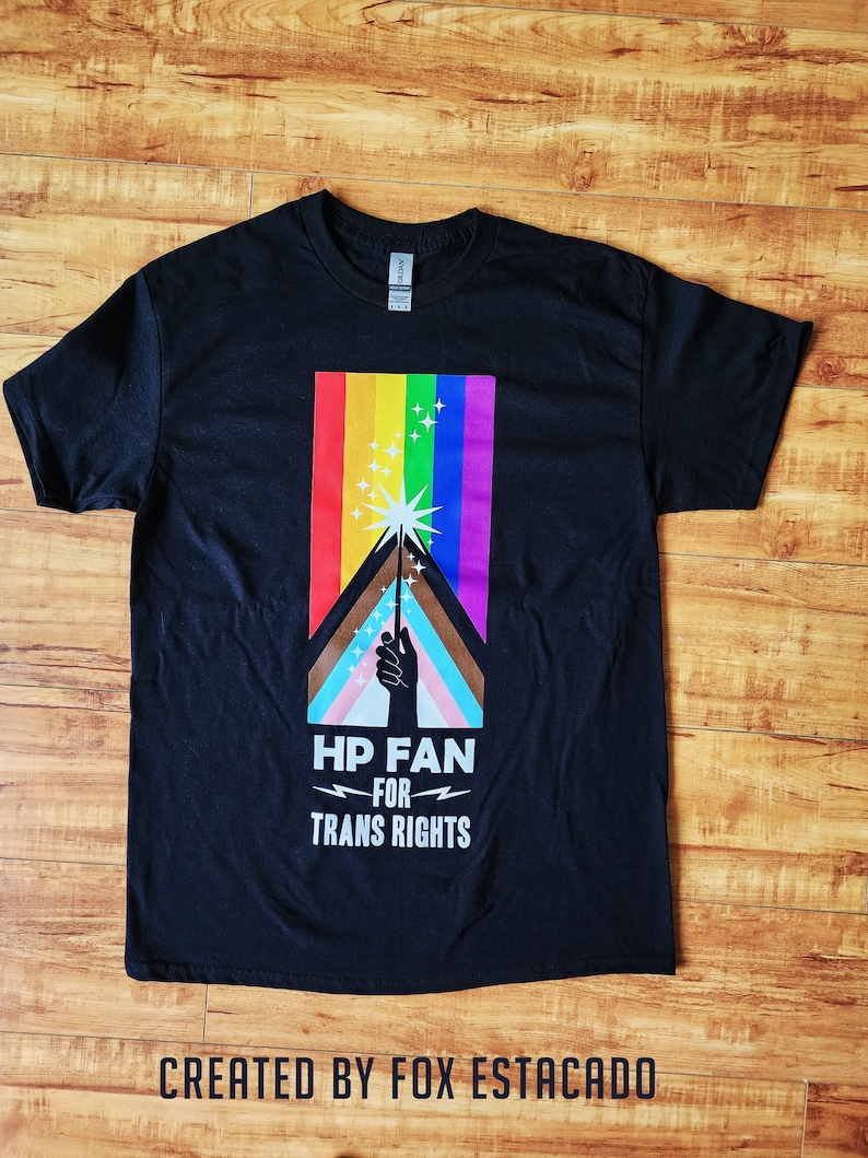 May include: Black t-shirt featuring a rainbow and a hand holding a wand. The text on the shirt reads "HP Fan for Trans Rights".