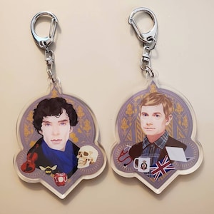 May include: Two keychains featuring illustrations of men. The keychain on the left features a man with dark hair and a blue scarf. The keychain on the right features a man with blonde hair and a plaid shirt. Both keychains have a purple background with gold accents.