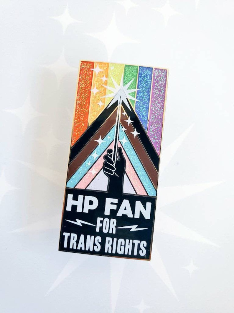 May include: A black enamel pin with a rainbow background and a hand holding a wand with a star on top. The text on the pin reads "HP FAN FOR TRANS RIGHTS".