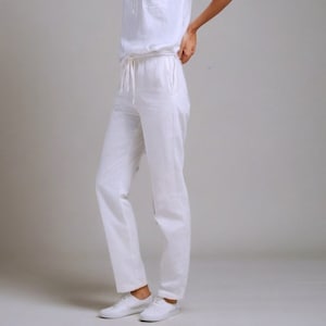 May include: A white linen jumpsuit with a v-neck top and a drawstring waist. The jumpsuit has a patch pocket on the left side of the chest.