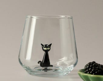 May include: A clear glass with a black glass cat figurine inside. The cat is sitting up and has a white face with black eyes and a small black mustache.
