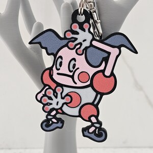 May include: A pink and gray cartoon character keychain with a silver key ring. The character has large eyes, a pink body, and gray wings.
