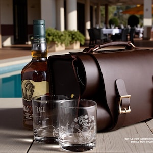 May include: A brown leather satchel with gold-colored buckles and a handle sits next to a bottle of whiskey and two clear glasses. The glasses have etched designs and the bottle has a label with a bison image.