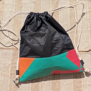Peut inclure: Sac à cordon noir avec un motif en blocs de couleurs. Le haut est noir, le bas présente des formes géométriques orange, vertes et rouges. Le sac a des cordons blancs en forme de corde.