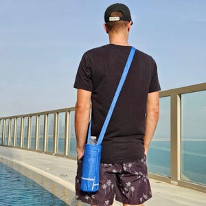 Könnte beinhalten: Ein blauer Wasserflaschenträger mit Schultergurt. Der Träger besteht aus einem strukturierten Material und hält eine transparente Wasserflasche. Die Person trägt ein schwarzes T-Shirt, schwarze Shorts und eine schwarze Baseballkappe.