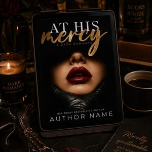 May include: A digital tablet displays the book cover "At His Mercy: A Dark Romance" with a close-up of a woman's lips. The image includes candles, a coffee cup, and a small book with the text "Morally questionable but make it Romance."