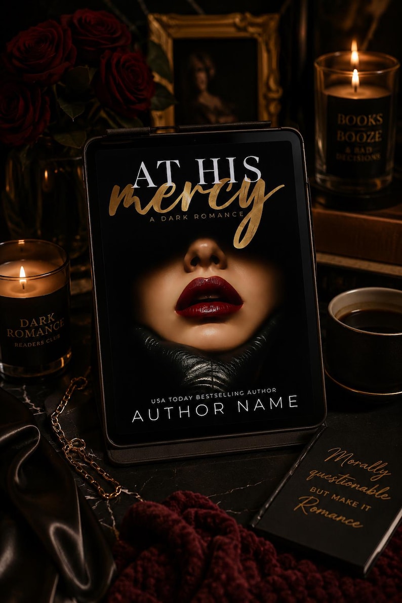 May include: A digital tablet displays the book cover "At His Mercy: A Dark Romance" with a close-up of a woman's lips. The image includes candles, a coffee cup, and a small book with the text "Morally questionable but make it Romance."